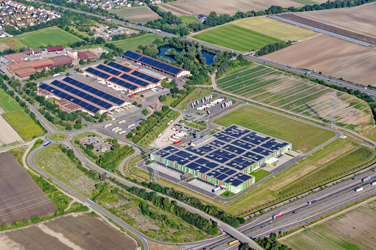 Das neue Vermarktungszentrum fällt mit seiner rot-grün gestreiften Fassage ziemlich auf, passt aber dennoch gut in die Landschaft. Es ist mittlerweile komplett mit Photovoltaikmodulen bedeckt und somit leistet die Genossenschaft einen bedeutenden Beitrag zur Reduzierung des CO
<sub>2</sub>
-Ausstoßes.